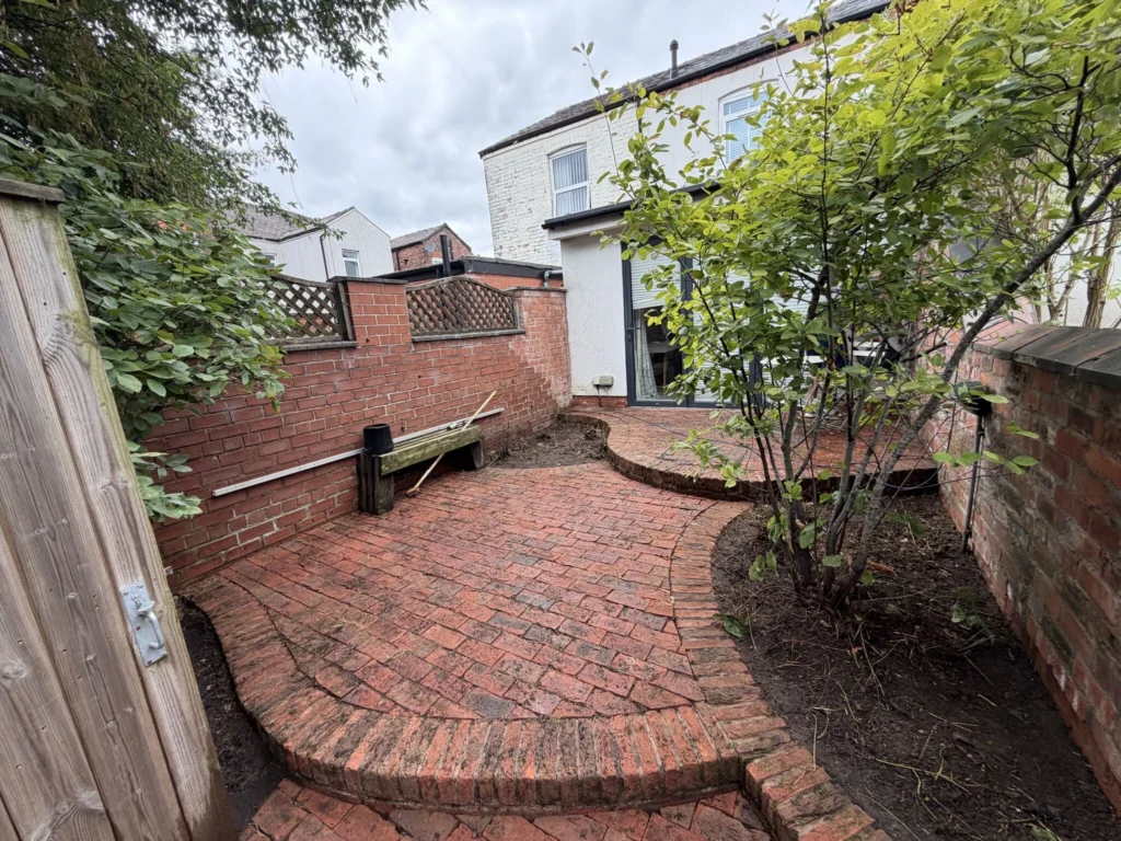 Brick-patio backyard with curved brick steps, brick walls, and green shrubs surrounding a white two-story house.