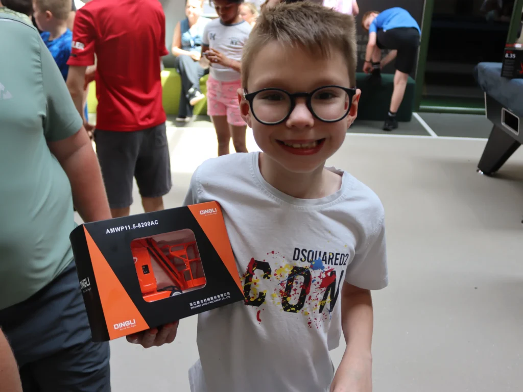 Smiling boy with glasses holding an orange-and-black boxed toy, indoors with people in the background.
