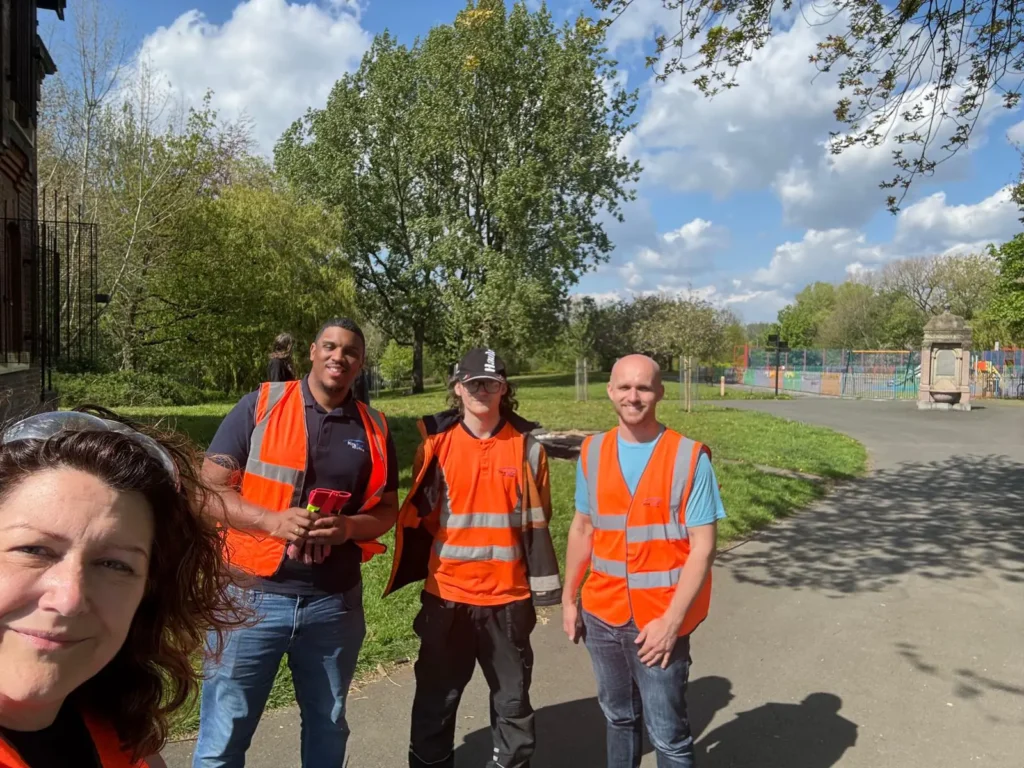 Four volunteers in high-visibility orange vests pose for a photo in a sunny park, with trees and a playground in the background.