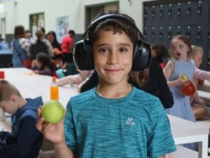 Smiling boy wearing large black headphones holds a green apple in a busy school cafeteria setting.