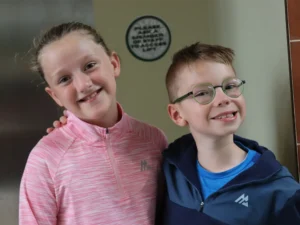 Two smiling children posing for a selfie indoors: girl in a pink striped jacket on the left and a boy with green glasses on the right.