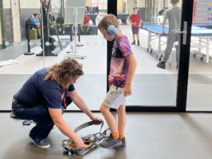 Adult kneeling on a gym floor helping a child buckle a harness or straps to a mobility device, with other kids watching in the background.