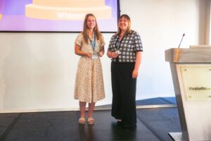 Two women stand on a stage; the left woman holds a glass trophy and both smile, with a large 'Winner' banner behind and a podium labeled 'thestudio' to the right.