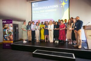 Group of award winners standing on a stage, posing with trophies during a ceremony, with a large screen in the background reading 'CONGRATULATIONS TO ALL OUR WINNERS!'