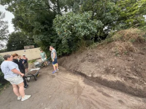 Group of people gathered beside a dirt path with wheelbarrows, listening to a man in shorts and a gray shirt under a leafy tree shade.