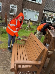 Woman in an orange safety jacket assembling a wooden bench in a garden with a brick house behind her.