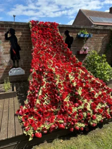 Red cascade of poppies forming a sculpture along a brick wall, flanked by two black silhouette cutouts and planter boxes, commemorating military service (Lifeworks sign nearby).