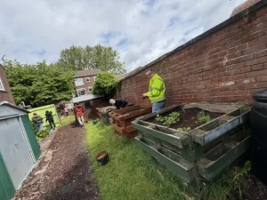 People gardening in a backyard with raised wooden planters along a brick wall; a man in a neon jacket is holding a tool.