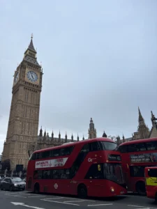 London skyline featuring the Elizabeth Tower (Big Ben) beside ornate palace architecture, with red double-decker buses in the foreground under a pale sky.