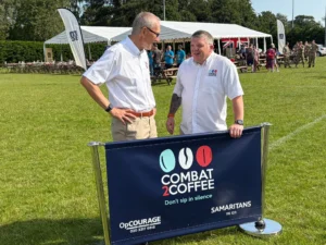 Two men in white shirts stand beside a banner for Combat Coffee at an outdoor event with tents and people in the background.