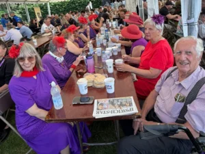 Crowd seated at long picnic tables under a white tent at an outdoor festival; many wear purple outfits and red hats pick up drinks and food.
