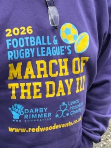 Close-up of a purple T-shirt with blue and yellow text announcing '2026 Football & Rugby League's March of the Day' and featuring logos for Darby Rimmer Foundation and Leeds Hospitals Charity with a website URL at the bottom.