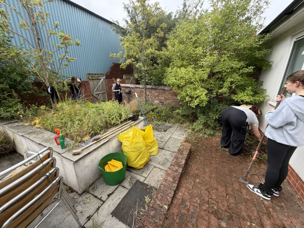 People gardening in a small backyard with tools, bags, and overgrown plants along a brick wall and blue metal building behind.