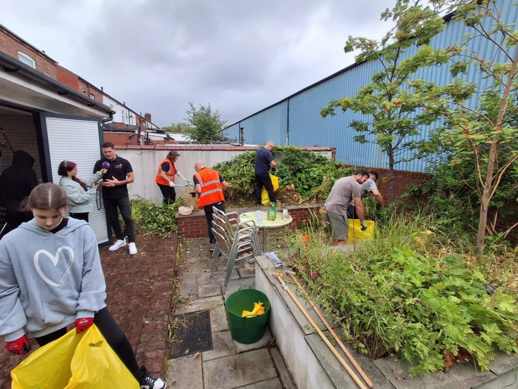 Group of volunteers cleaning a backyard garden, with orange vests, yellow trash bags and gardening tools nearby.