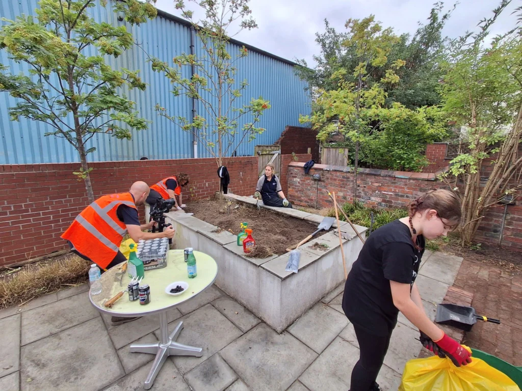 Group of volunteers in orange vests working in a brick-walled garden with tools and a camera on a round table in the foreground.