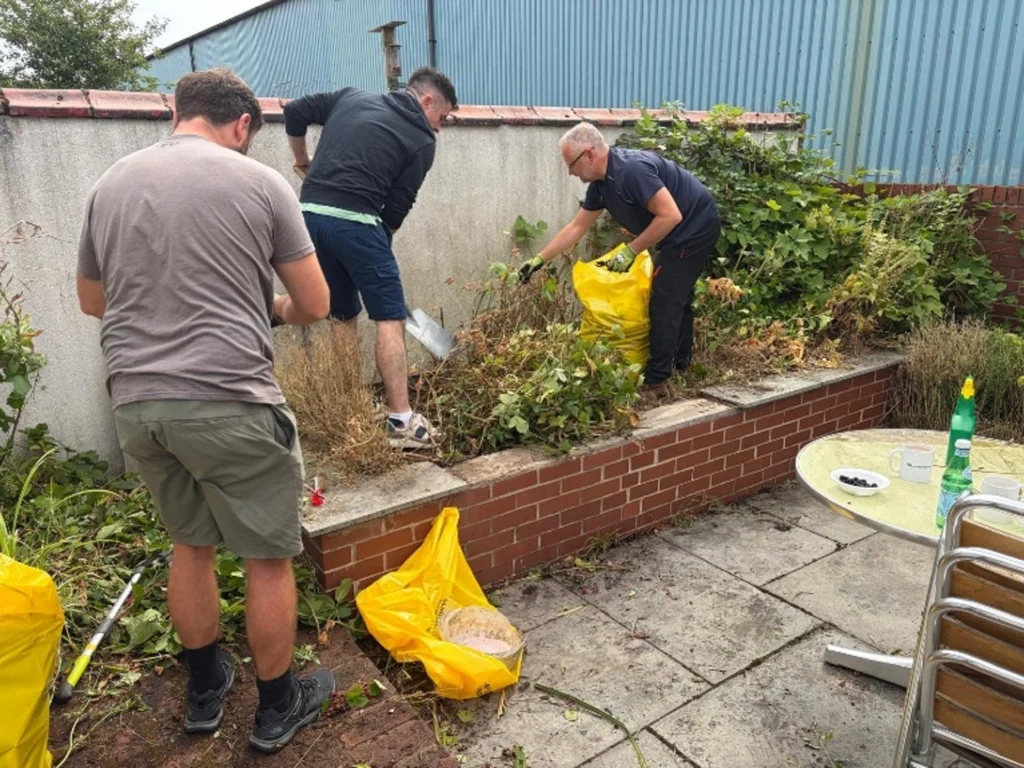 People gardening in a small backyard with tools, bags, and overgrown plants along a brick wall and blue metal building behind.