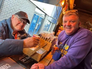 Two men at a cafe counter smiling; the older man in a cap holds a cup while the other man in a purple sweatshirt holds a mug behind a wooden display of snacks.