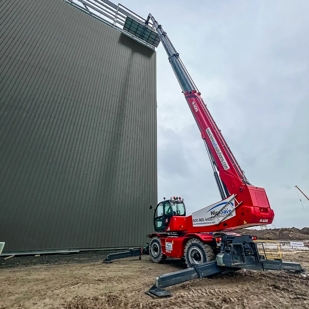 Red mobile crane with its boom extended, lifting a platform to the top edge of a tall dark-green metal wall at a construction site. The crane is stabilized on wide outriggers on dirt ground.
