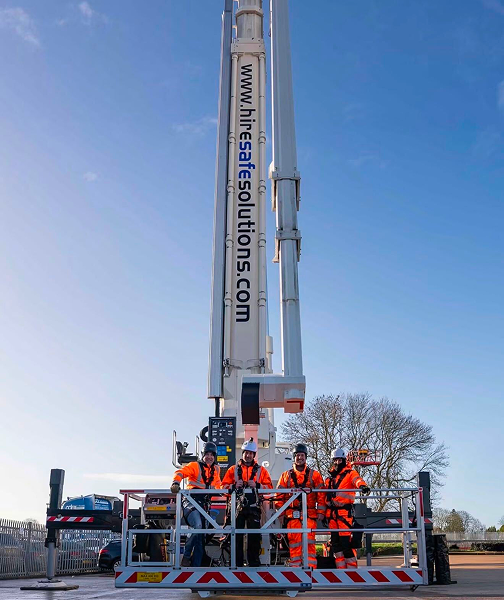 Four workers in orange high-visibility suits and hard hats stand on a raised work platform attached to a tall hydraulic crane, with the site’s banner www.hifesolutions.com visible against a clear blue sky.