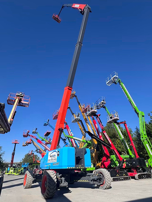 Blue sky above a row of colorful elevated work platforms (boom lifts) with their arms raised on a paved lot, showcasing equipment at a dealership or rental yard.