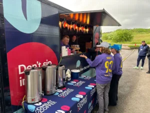Mobile coffee trailer with warm string lights serving customers at an outdoor event; volunteers in purple sweatshirts (number 93) take orders.