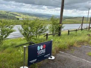 Blue Combat Coffee banner set up along a rural roadside with green fields and hills in the distance.