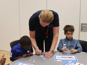 Adult man guiding two boys as they sign their pictures with markers at a classroom-style table.