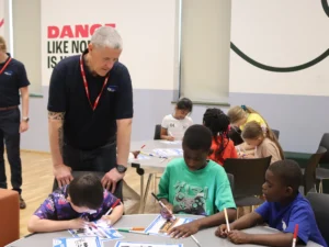 Group of children at a table coloring worksheets while two adults supervise in a bright classroom gym.