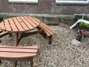 Round brown wooden picnic table on a gravel surface beside a brick wall, with a wheelbarrow holding plants on the right.