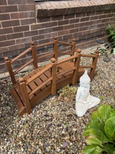 Brown wooden garden bench frame and railing pieces set on a gravel yard beside a brick wall, with a white Buddha statue nearby.