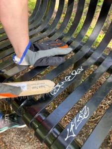 Gloved hand pressing a wire brush against a curved black metal park bench outdoors, removing dirt from graffiti-marked slats.