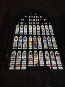 Tall Gothic stained-glass window with numerous heraldic crests and colorful panels inside a dim stone cathedral.