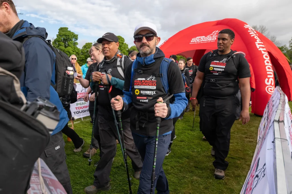 Group of hikers in black shirts with backpacks at a charity walk start line under a red archway reading 'Walking With The Wounded'