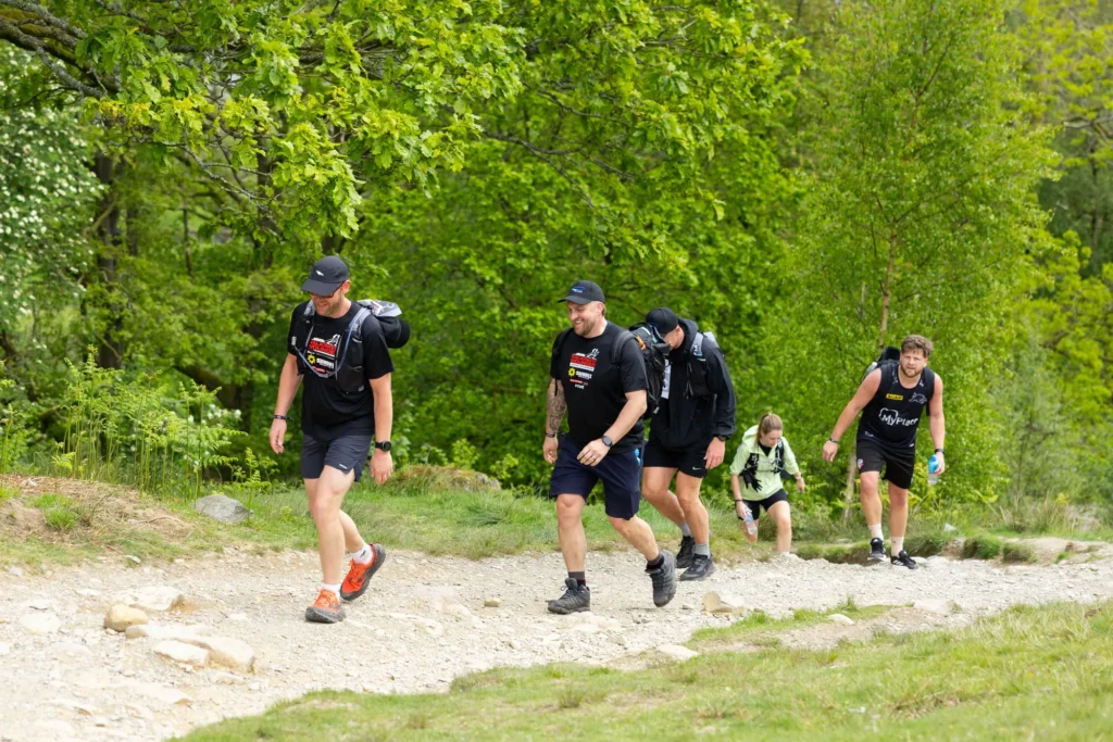 Group of hikers with backpacks walking on a rocky trail through a lush green forest, smiling in the sunshine.
