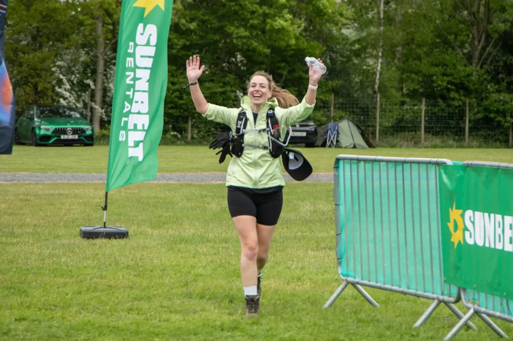 Female runner finishes a race with arms raised in celebration beside green event banners on a grassy field.
