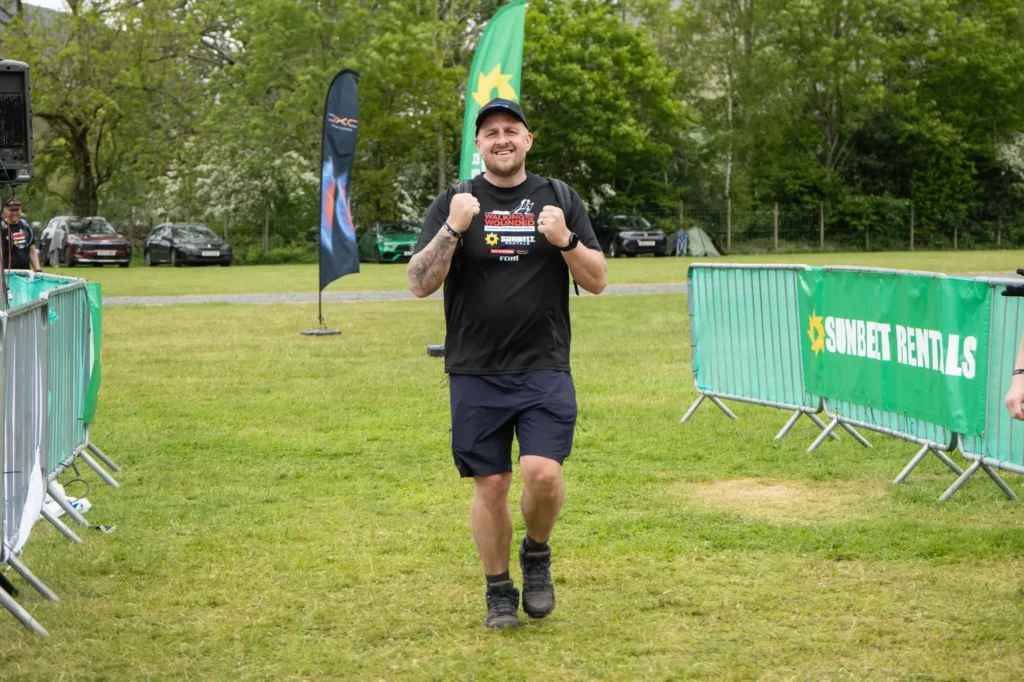 Smiling man in black t-shirt and cap runs toward the camera at an outdoor event, with green banners and crowd barriers in the background.