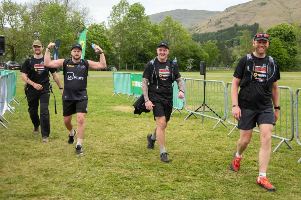 Four men in black athletic shirts with packs walk a grassy course, barriers and green banners visible, hills in the background.