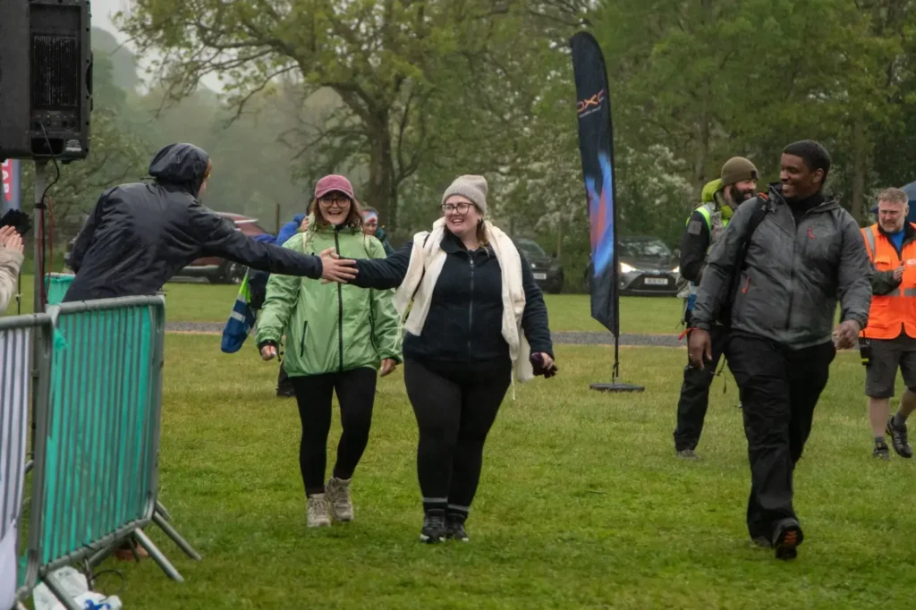 Two women walk toward the camera and shake hands with a man reaching from the left, as event volunteers in hi-vis jackets stand in the background.